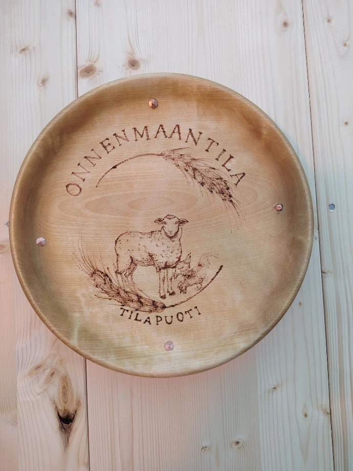 Onnenmaan tilapuodin puinen kyltti, johon on poltettu tilan logo – tilan isännän valmistama ja tyttären viimeistelemä käsityö.